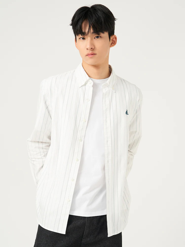 Male model wearing the Beanpole Men Oxford Stripe Shirt in White, styled with a black tie and beige trousers while holding a red apple. The shirt is crafted from 100% cotton with a comfort fit, offering timeless versatility for both workwear and casual looks.