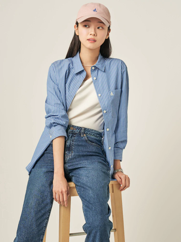 Female model seated in the Beanpole Ladies Blue Stripe Semi-Overfit Shirt in Sky Blue, styled over a white tank and paired with mid-wash denim. The shirt is made from a cotton–polyester blend and features a semi-overfit cut designed for versatile, everyday styling.