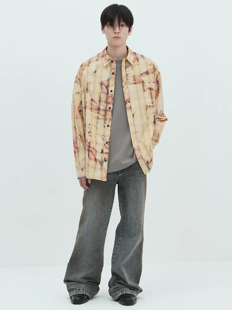 Male model wearing the JUUN.J Sunbleach Printed Check Shirt in Yellowish Brown, styled open over a gray T-shirt with wide-leg faded jeans. The shirt is made from 100% cotton and features a loose fit with distressed print detailing for a contemporary edge.