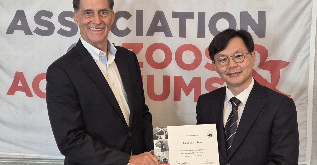 Two representatives stand in front of an Association of Zoos and Aquariums banner, holding and displaying Everland Zoo’s AZA reaccreditation certificate while shaking hands and smiling for the camera.