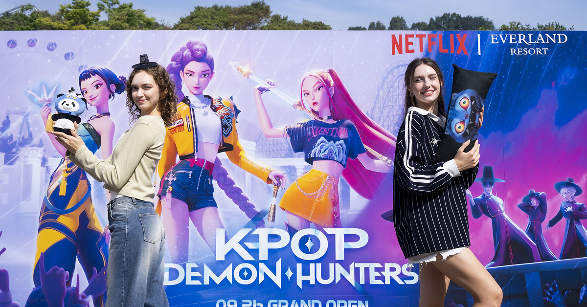 Two visitors holding themed merchandise in front of a large KPop Demon Hunters backdrop at Everland.