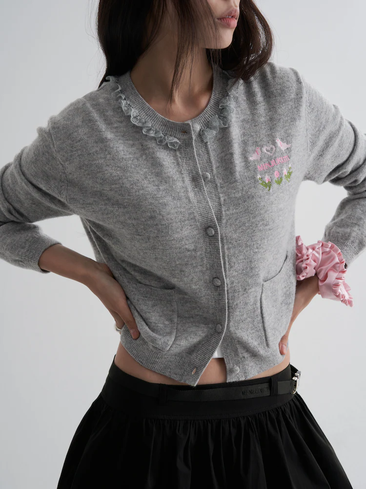 The 8seconds MINJUKIM Neck Ruffle Cardigan in Grey, detailed with front buttons, patch pockets, and floral embroidery on the chest. The design includes a ruffled neckline and is paired with a pink satin scrunchie, emphasizing whimsical elegance in a casual silhouette.