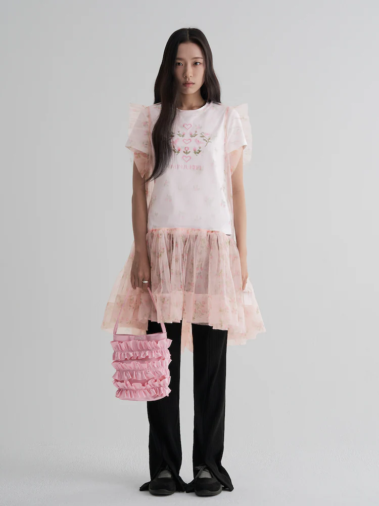 The 8seconds MINJUKIM Flower Pattern Sleeveless Dress in Light Pink showcases a lightweight tulle overlay adorned with delicate floral prints. Styled over black pants and paired with a ruffled pink tote, the look blends whimsical detailing with modern layering.