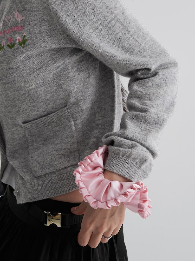 The 8seconds MINJUKIM Satin Ruffle Scrunchie in Light Pink, styled on the wrist over a grey cardigan sleeve. The accessory features a lustrous satin texture and layered ruffles, bringing a whimsical touch to casual outfits.