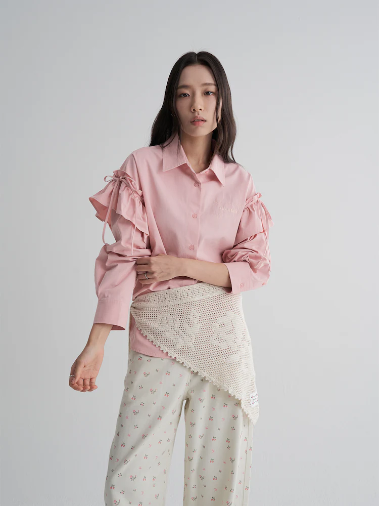 The 8seconds MINJUKIM Frill Long-Sleeve Shirt in Light Pink, detailed with cascading ruffles and adjustable ribbon ties along the sleeves. Styled with a crochet wrap detail and printed skirt, the shirt brings a feminine twist to structured tailoring.