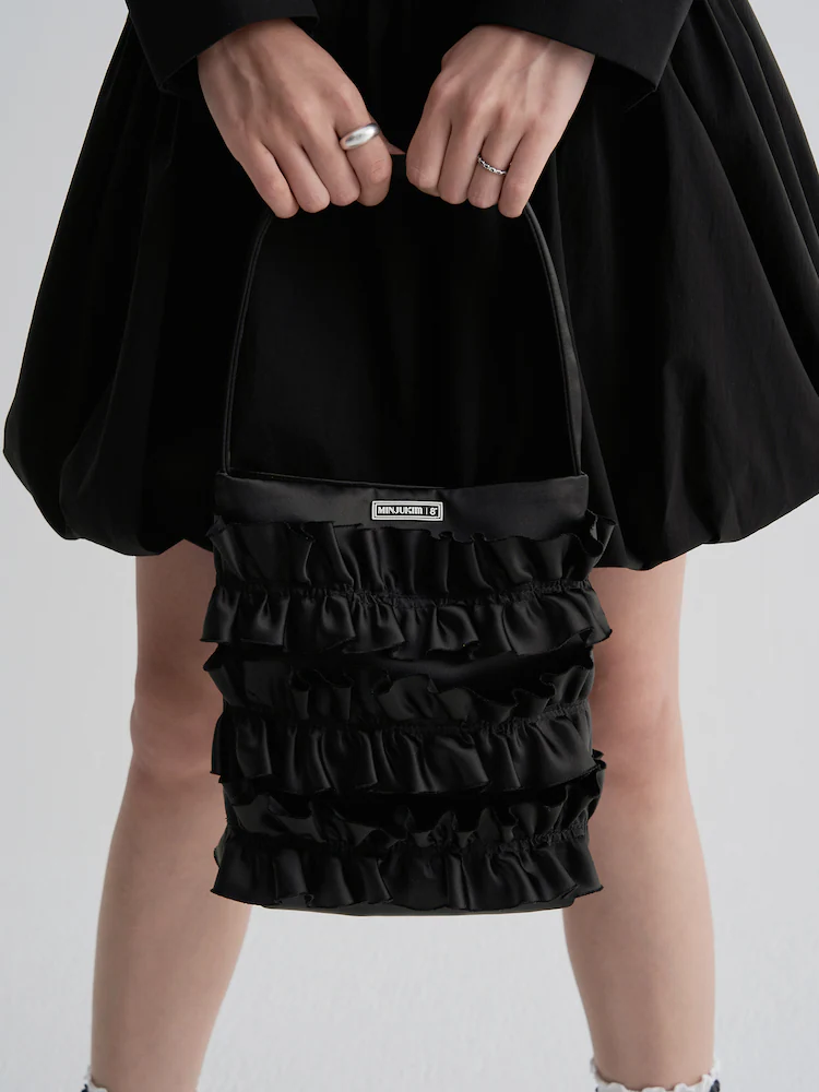The 8seconds MINJUKIM Shirring Mini Tote Bag in Black, designed with multiple horizontal ruffle tiers and a slim shoulder strap. Finished with a MINJUKIM logo plate, the bag offers a whimsical yet versatile accent to the collection’s casual-romantic looks.