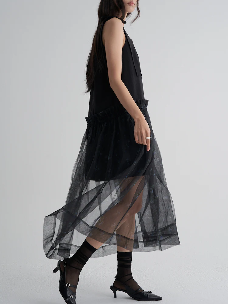 The 8seconds MINJUKIM Sheer Long Frill Dress in Black, designed with a structured underdress and a transparent tulle overlay. The lightweight fabric is softly pleated, revealing delicate floral accents while adding depth and movement.