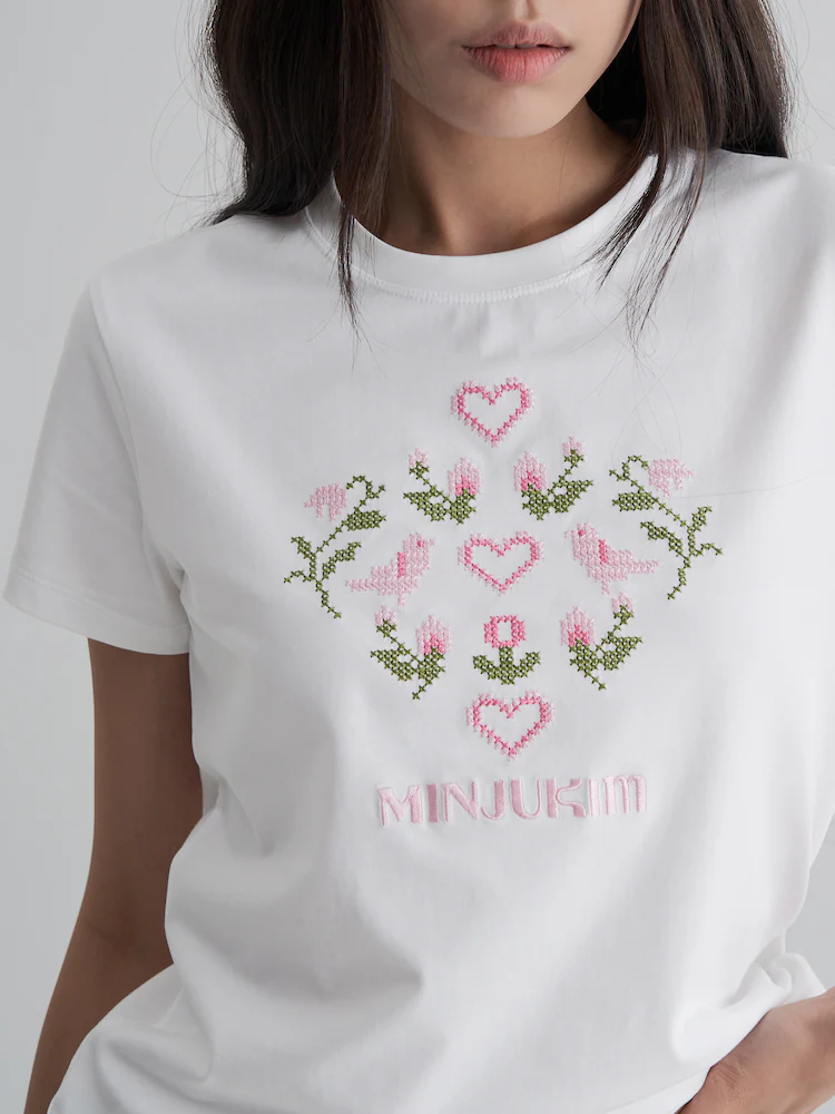 The 8seconds MINJUKIM Graphic Slim-Fit T-Shirt in Ivory, designed with pink cross-stitch embroidery of flowers and hearts on the chest. The slim silhouette and soft cotton fabric make it a versatile everyday piece with whimsical charm.