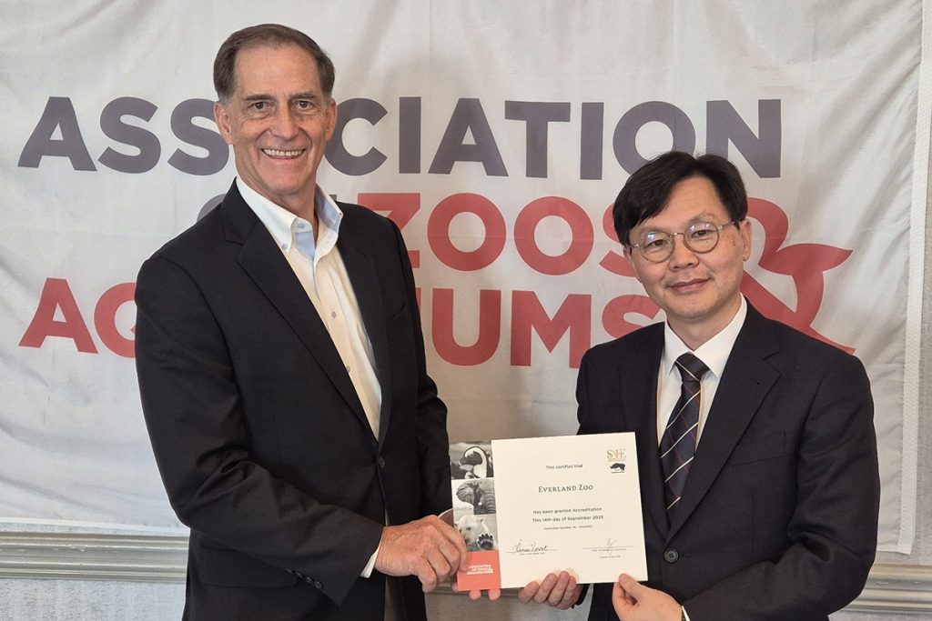 Two representatives stand in front of an Association of Zoos and Aquariums banner, holding and displaying Everland Zoo’s AZA reaccreditation certificate while shaking hands and smiling for the camera.