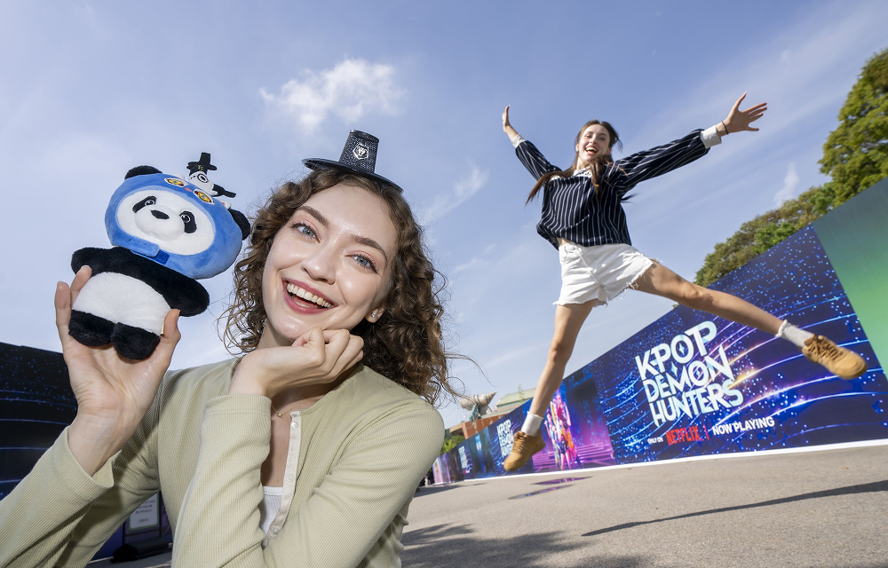 A visitor holding a panda plush toy while another jumps in front of a KPop Demon Hunters backdrop at Everland.