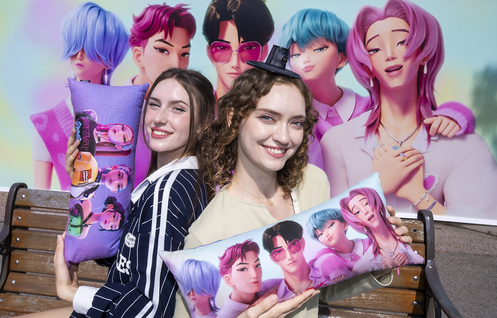 Two visitors holding colorful KPop Demon Hunters character cushions in front of a themed backdrop at Everland.