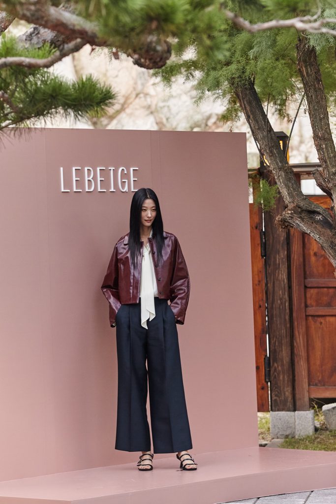 Model Park Ji-hye poses in front of the LEBEIGE logo backdrop at Gala de LEBEIGE, wearing a burgundy jacket, white blouse, and wide black trousers.