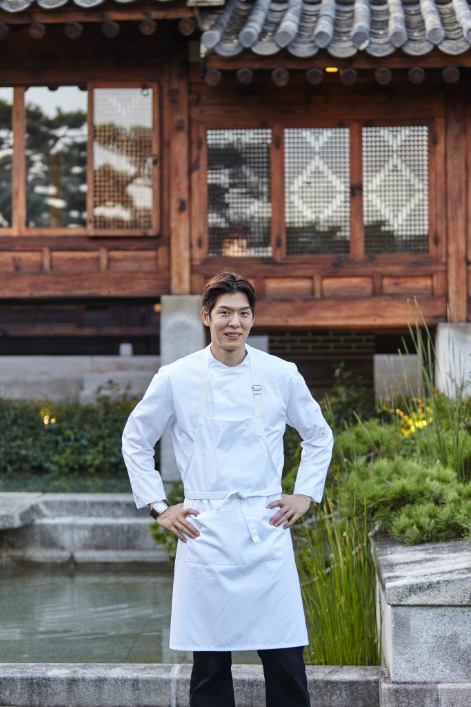Chef Austin Kang stands outdoors in front of the Korea Furniture Museum, wearing a white chef’s uniform for his role at Gala de LEBEIGE.