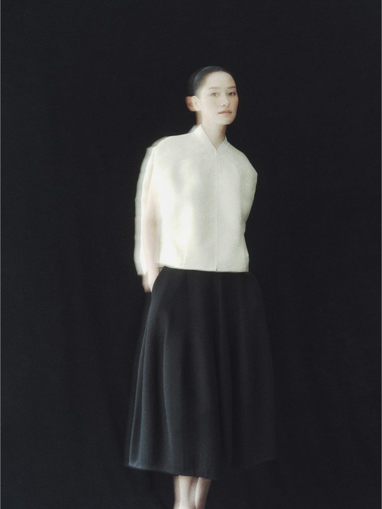 A model poses against a dark backdrop wearing a white structured top and a flowing black skirt from the LEBEIGE × Heechan Kim capsule collection.