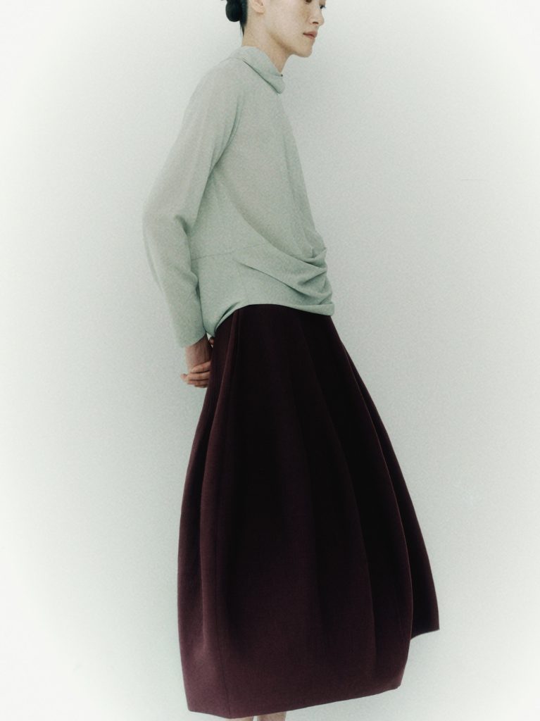 A model in a light green top and voluminous burgundy skirt poses against a pale background, showcasing a piece from the LEBEIGE × Heechan Kim capsule collection.