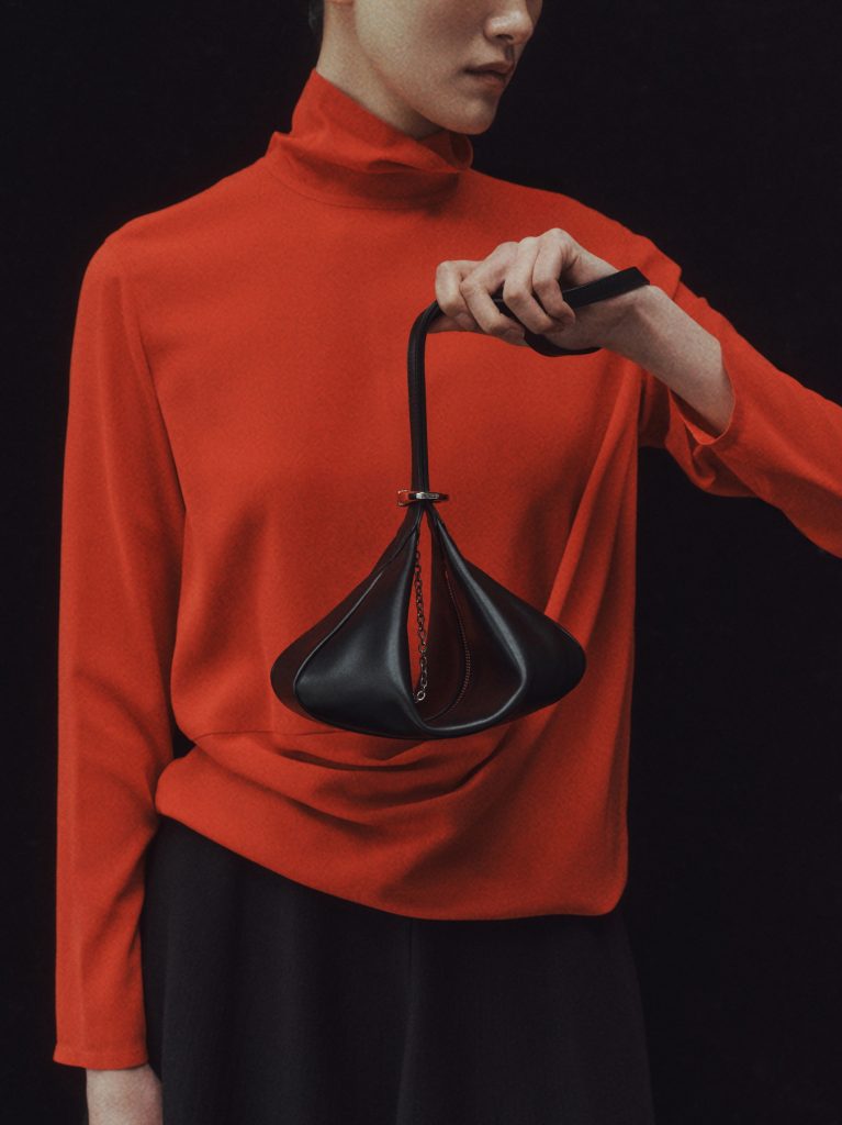 Close-up of a model in a red top holding a black leather bag with a sculptural shape, part of the LEBEIGE × Heechan Kim collaboration.
