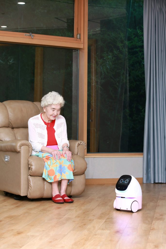 An elderly woman sitting in a chair smiles at a small AI companion robot on the floor, designed to assist with daily life and provide interaction.