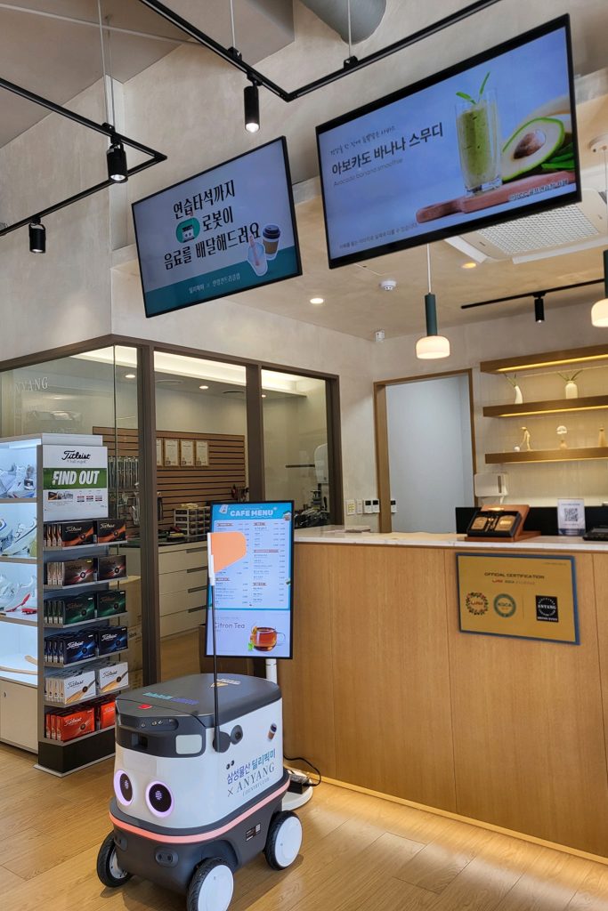 A small delivery robot stands inside a café beneath digital menu screens, equipped to transport drinks to customers’ tables.