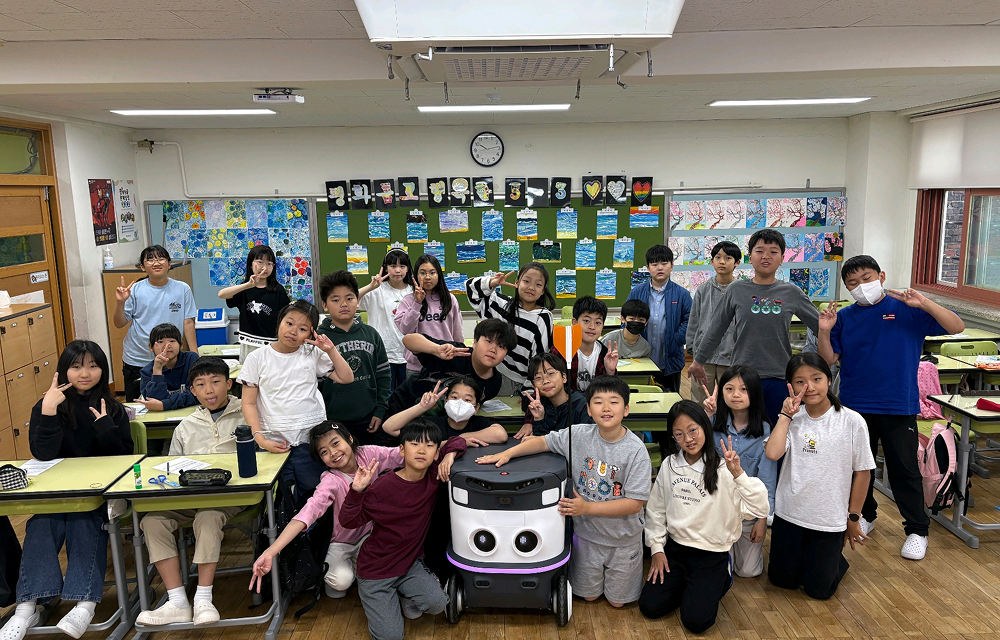A group of students pose around a small delivery robot in a classroom, highlighting its educational use in showing future robotics applications.