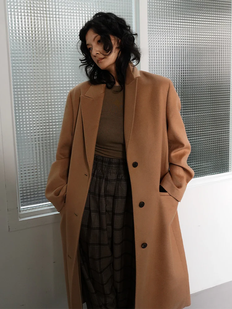 Model wearing a long camel-brown wool coat over a brown knit and check skirt, styled with KUHO’s elegant tailoring.