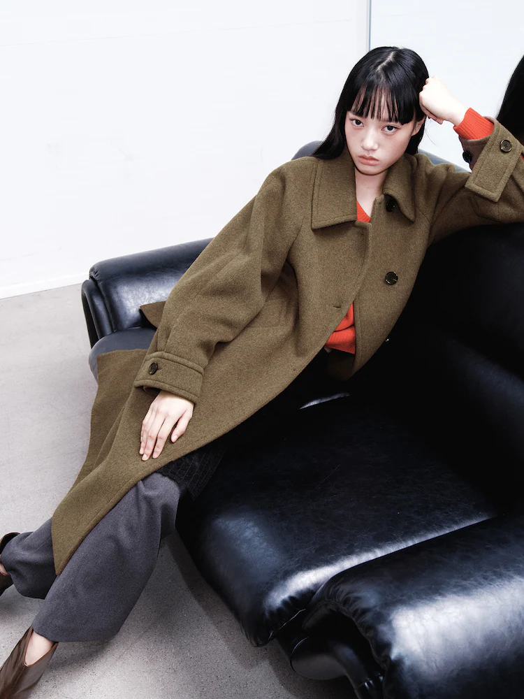 Model reclining on a black leather sofa wearing a yellowish brown wool coat and red knit top, representing kuho plus’s modern volume styling.