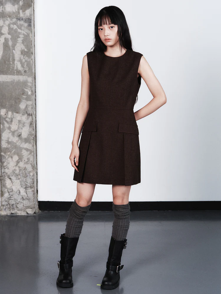 Model standing in a minimalist setting wearing a dark brown sleeveless dress with pockets and gray socks, highlighting kuho plus’s structured minimalism.