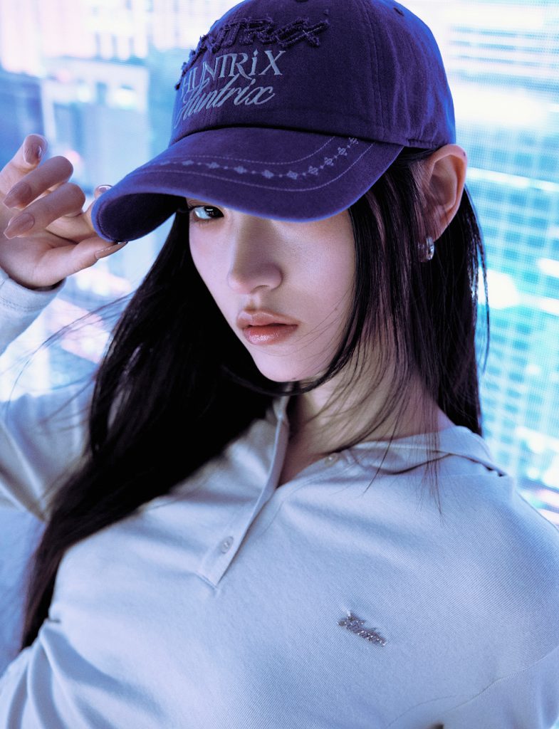 Close-up of a model wearing a purple baseball cap embroidered with the word “Huntrix” from the 8seconds x K-Pop Demon Hunters collection. She pairs it with a white collared top and silver earrings, posing against a city-inspired digital backdrop.