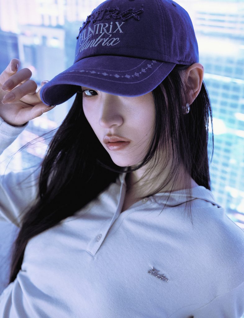 Close-up of a model wearing a purple baseball cap embroidered with the word “Huntrix” from the 8seconds x K-Pop Demon Hunters collection. She pairs it with a white collared top and silver earrings, posing against a city-inspired digital backdrop.