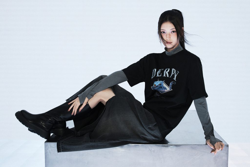 Female model sitting on a metallic platform wearing a black “Derpy” T-shirt from the 8seconds x K-Pop Demon Hunters collection. The outfit is paired with a gray long-sleeve inner top, a long dark denim skirt, and black knee-high boots against a minimalist light background.