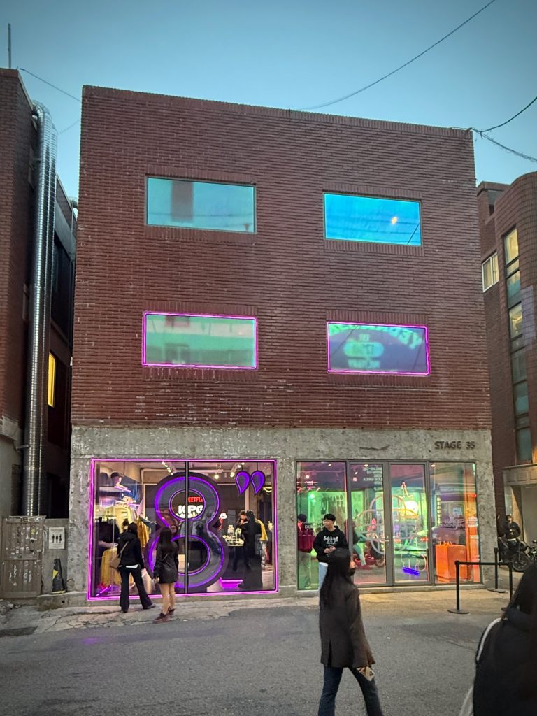A three-story brick building housing the 8seconds x K-Pop Demon Hunters pop-up store at Stage35 in Seongsu. The storefront features purple neon lighting and large display windows showing collaboration merchandise and decorations inside.