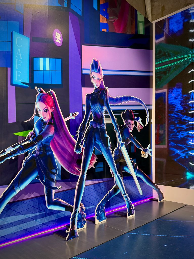 Display installation inside the 8seconds x K-Pop Demon Hunters pop-up store showing three full-size standees of “Huntrix” characters posed in dynamic battle stances against a backdrop resembling a cityscape from the film.