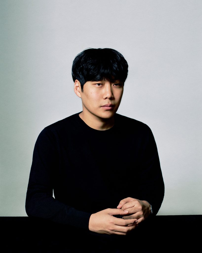 Portrait of designer Jaden Cho sitting against a light gray background, wearing a black sweater with hands resting in front.