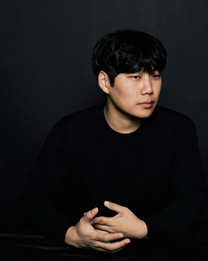 Portrait of designer Jaden Cho sitting against a dark background, wearing a black sweater with hands folded.