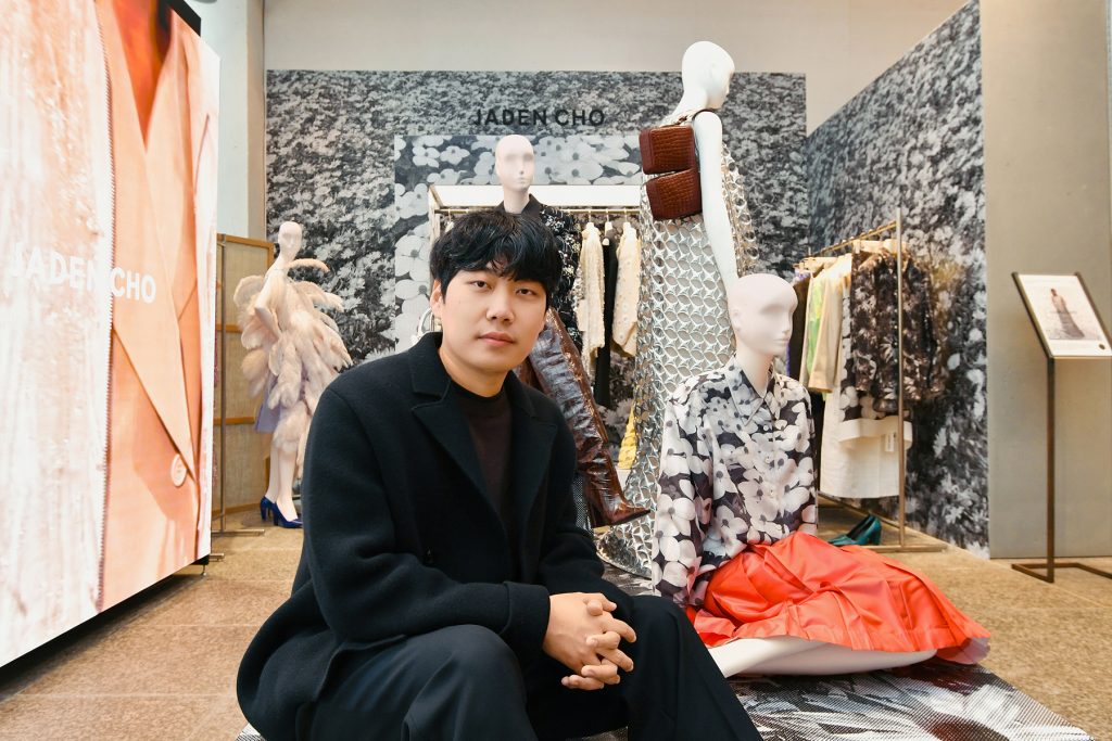 Designer Jaden Cho seated inside the BEAKER Cheongdam exhibition space, surrounded by mannequins dressed in pieces from the JADEN CHO collection, including floral prints and textured dresses.