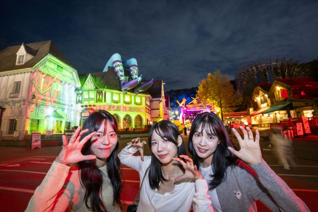Three visitors posing at night in front of Everland’s Blood City area, surrounded by colorful illuminated buildings and a giant witch’s leg sculpture on the roof.