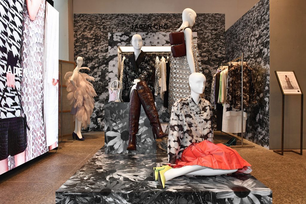 Mannequins showcasing JADEN CHO pieces, including a textured silver dress and floral-patterned looks, inside the BEAKER Cheongdam exhibition space with racks of clothing in the background.
