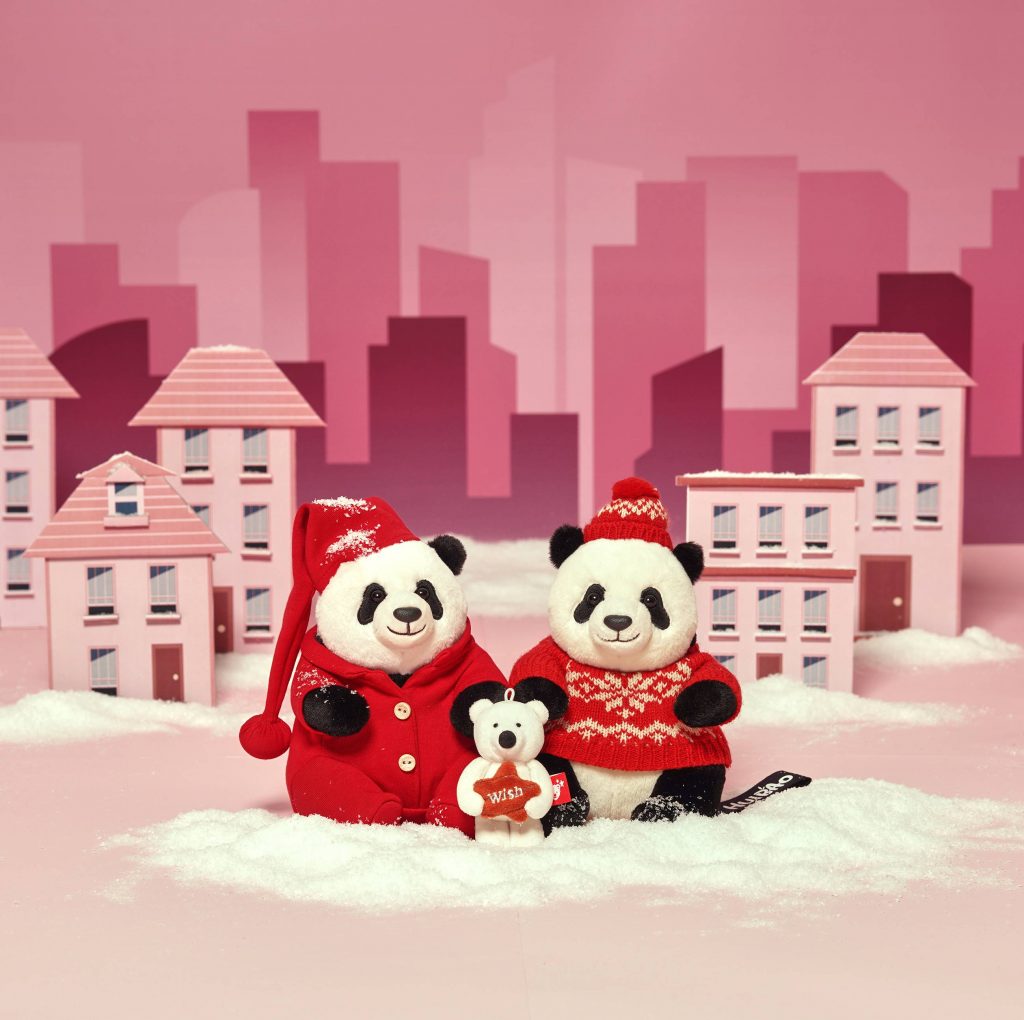 Two panda plush toys dressed in red winter clothing, Rui Bao and Hui Bao, posed in a snowy scene with miniature pink houses, joined by a small polar bear holding a “wish” heart.