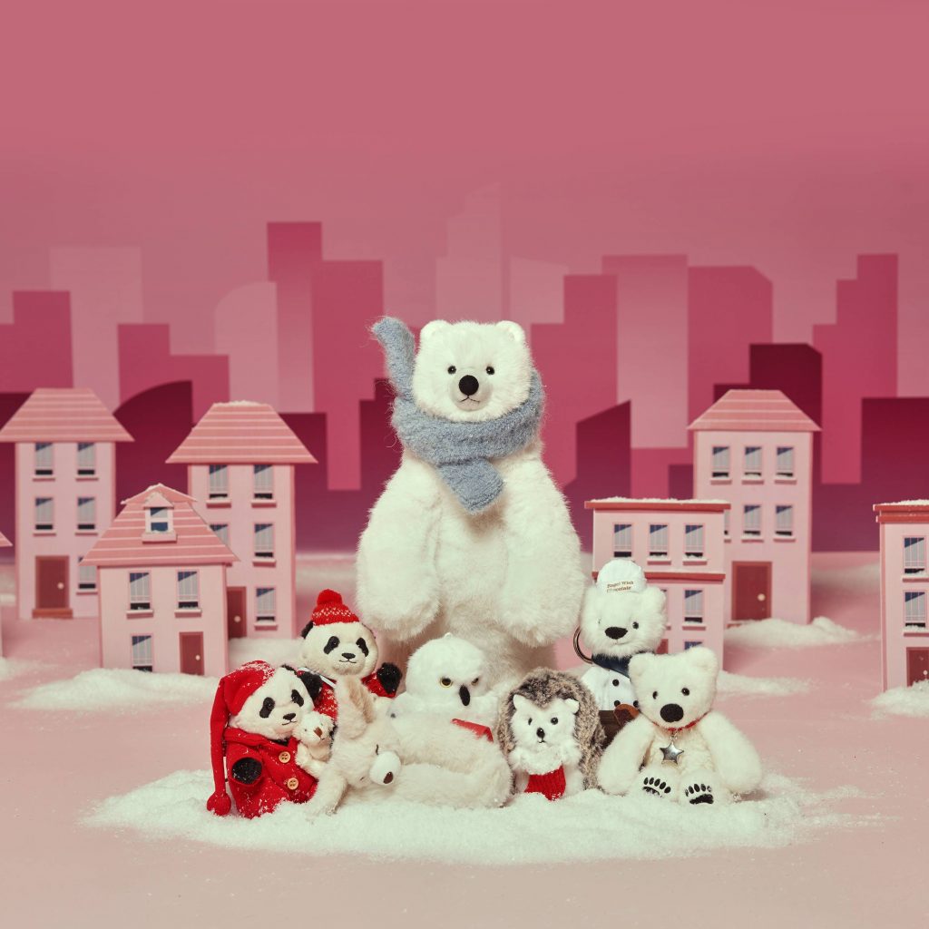 Assorted Bagel Wish Bear plush toys arranged in a snowy display against a pink village backdrop, featuring a large polar bear with a scarf, a chef bear, a small polar bear, a hedgehog, a white owl, and additional winter animal characters.