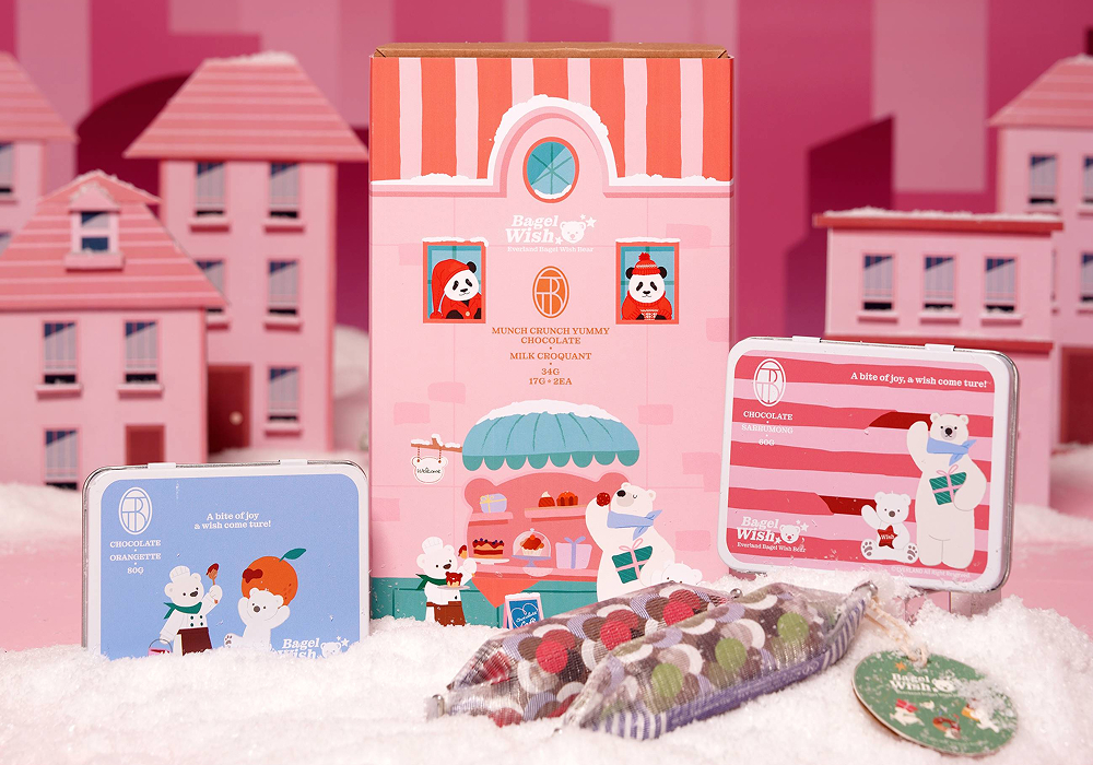 A display of Bagel Wish Bear x TREETOBAR collaborative chocolate products, including illustrated tin boxes and a gift set, arranged on snow with a pastel pink village backdrop.