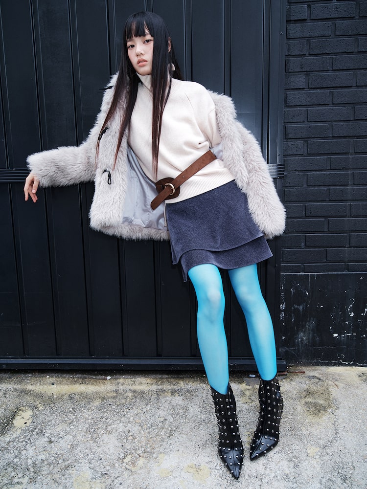 Model wearing a navy velvet corduroy cancan mini skirt styled with a cream turtleneck sweater, faux-fur jacket, and bright blue tights against a black brick backdrop.