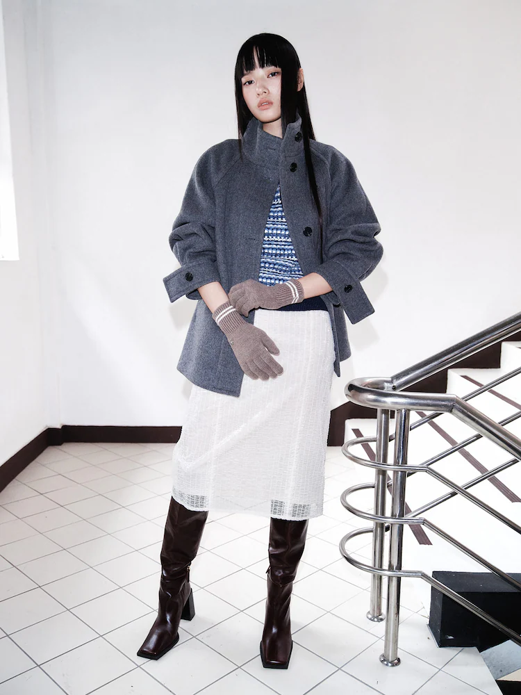 Model wearing an ivory layered skirt with a velvet underlay, styled with a gray jacket, knit gloves, and tall brown boots on an indoor staircase backdrop.