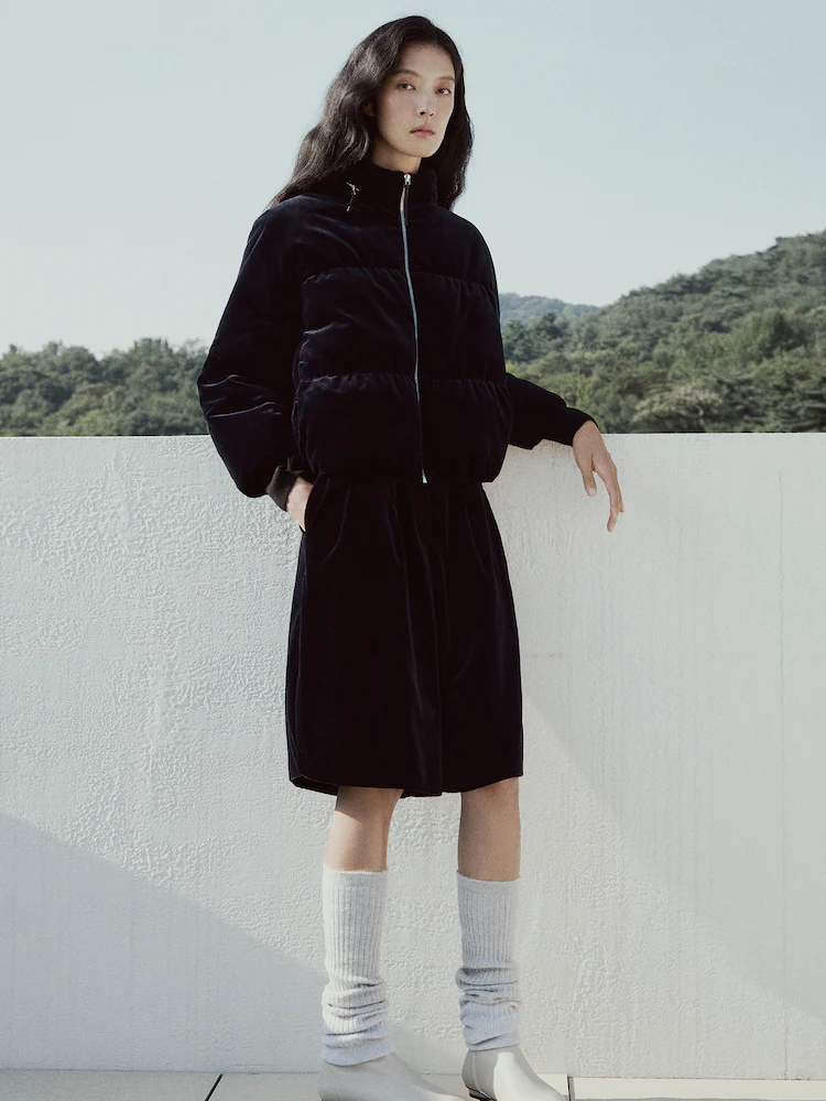 Model wearing a navy velvet cropped puffer down jumper, standing outdoors against a light concrete railing with trees in the background.