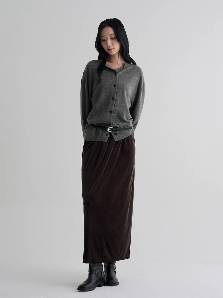 Model wearing an ash-toned velvet maxi skirt styled with a grey buttoned cardigan and black ankle boots, photographed against a minimal light grey studio backdrop.