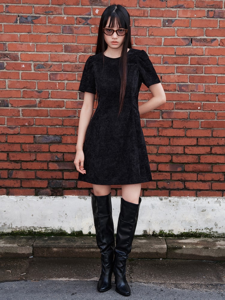 Model wearing a black velvet mini dress styled with tall black boots, standing against a red brick wall outdoors.