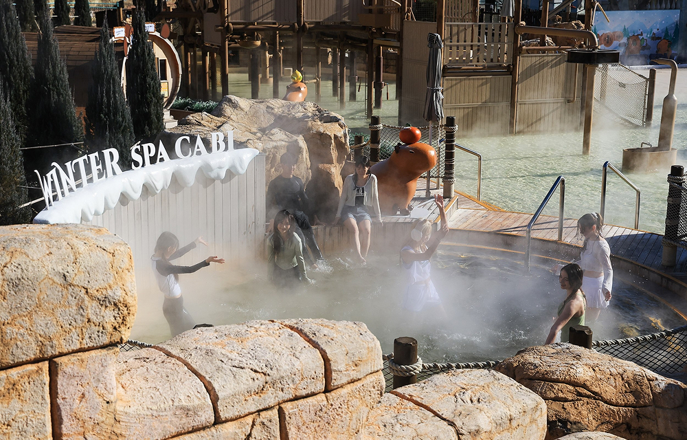 Visitors enjoy a steaming outdoor spa pool at Caribbean Bay’s Winter Spa Cabi, with rocky landscaping, winter-themed signage, and surrounding attractions visible in the background.