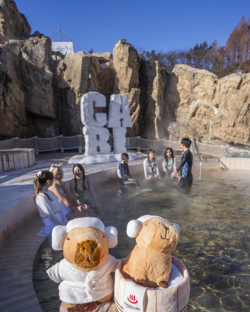 Visitors sit in a steaming outdoor spa pool at Caribbean Bay’s Winter Spa Cabi, with large “CABI” signage and rocky cliff structures in the background, along with capybara plush toys in the foreground.