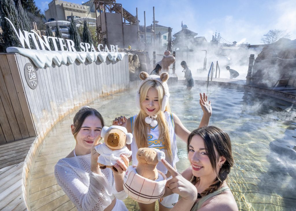 Three guests pose with capybara plush toys while relaxing in a steaming outdoor pool at Caribbean Bay’s Winter Spa Cabi, with winter-themed signage and spa facilities visible in the background.