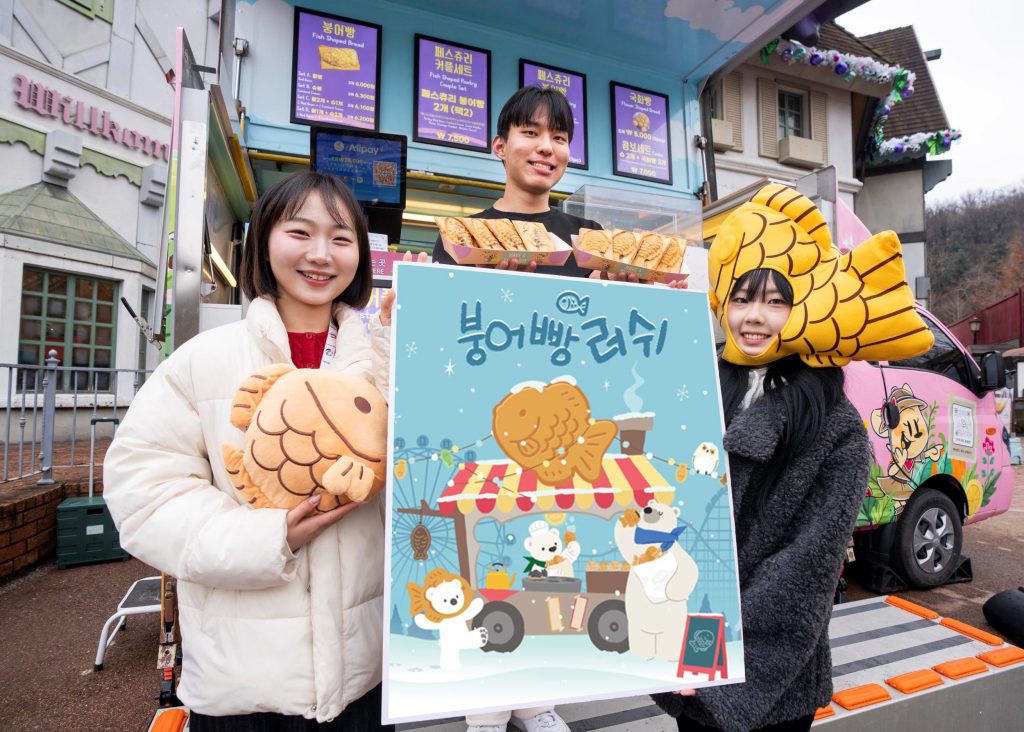 Three models holding an illustrated “Bungeoppang Rush” poster in front of a winter-themed food truck selling fish-shaped pastries at Everland