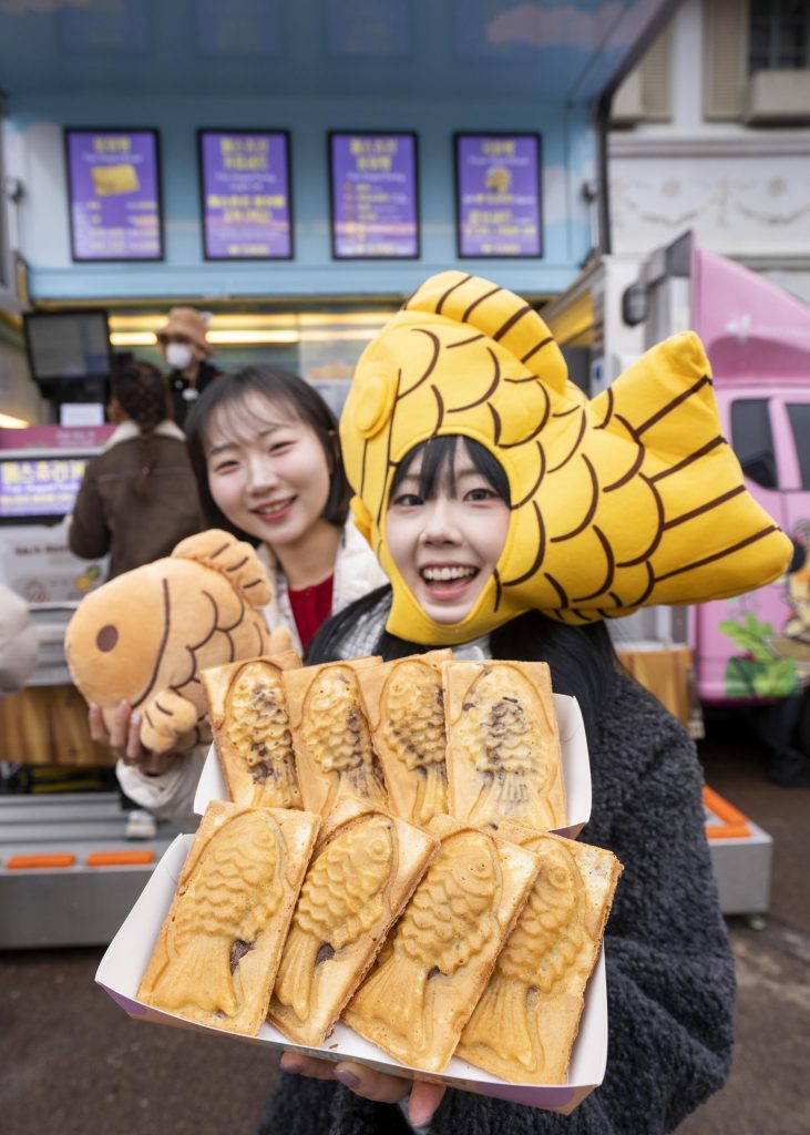 Smiling visitors holding multiple bungeoppang pastries in front of a winter-themed Everland food booth