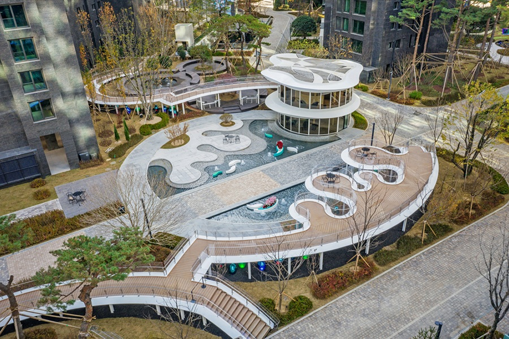 Aerial view of Blue Forest Garden at Jamsil Raemian I’Park featuring curved walkways, water features, seating areas, and landscaped greenery between residential buildings.